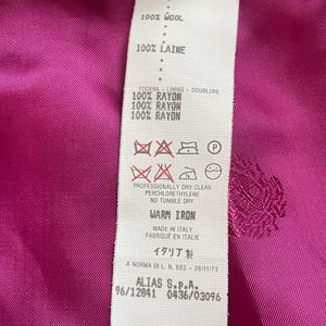 May include: A vibrant magenta garment interior featuring a white fabric care label. The label specifies 100% wool composition with a 100% rayon lining, includes care symbols, states professionally dry clean, no tumble dry, and warm iron, made in Italy.