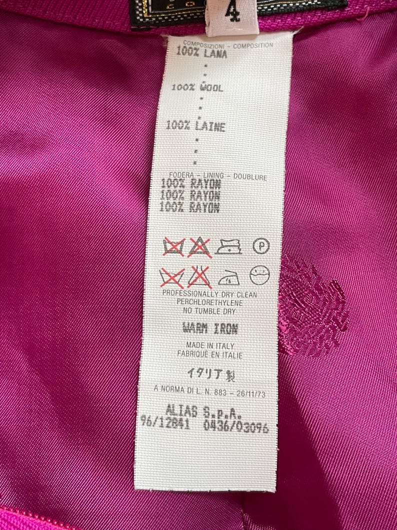May include: A vibrant magenta garment interior featuring a white fabric care label. The label specifies 100% wool composition with a 100% rayon lining, includes care symbols, states professionally dry clean, no tumble dry, and warm iron, made in Italy.