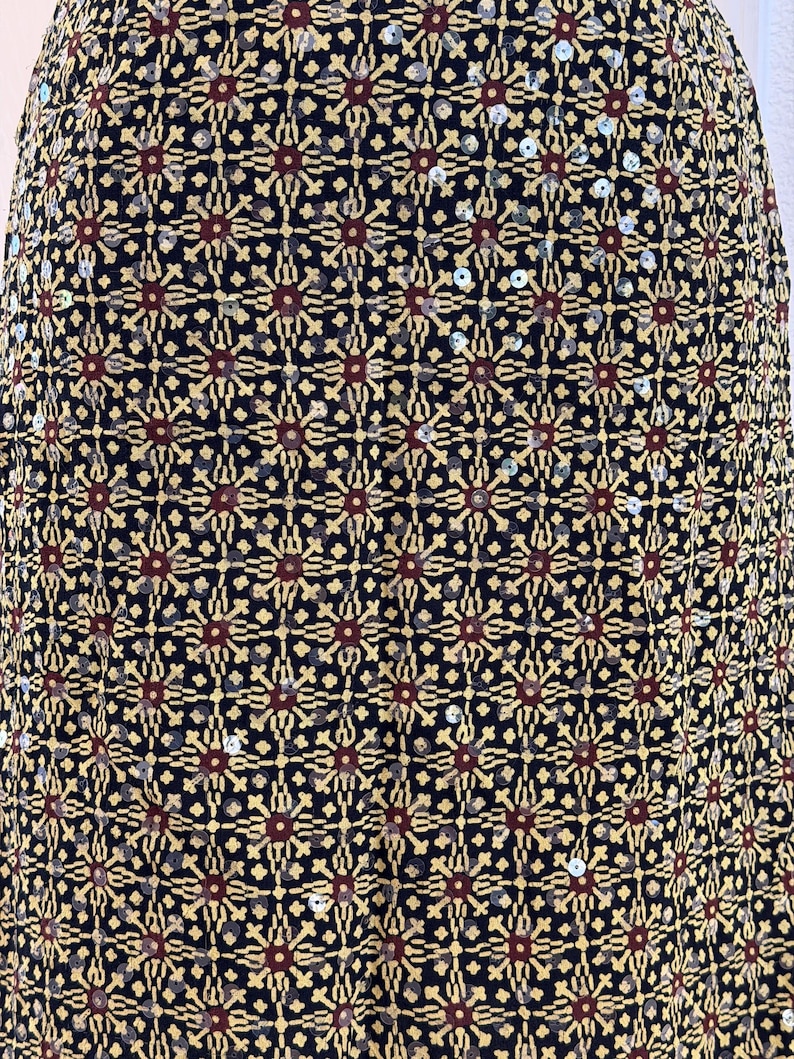 May include: A black fabric skirt featuring a repeating geometric pattern in gold and dark red. The design consists of star-like motifs arranged in a grid, accented with small, reflective silver sequins scattered throughout the entire surface.