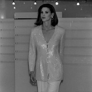 May include: A long-sleeved blazer featuring a V-neckline and a front button closure. The garment is covered in a dense, reflective sequin texture that catches the light, creating a shimmering appearance. It is paired with plain, light-colored trousers.