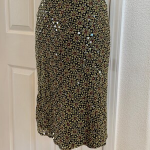 May include: A knee-length skirt featuring a black base with a repeating geometric pattern in shades of yellow, red, and green. The fabric is embellished with small, reflective silver sequins scattered throughout the intricate design.