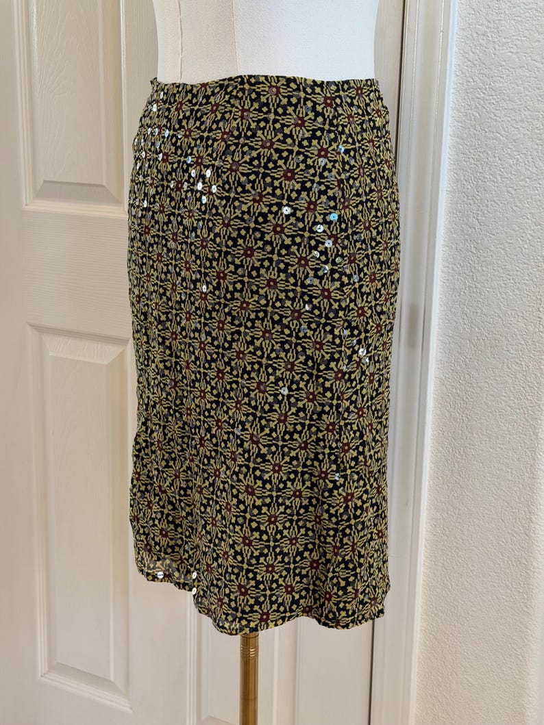 May include: A knee-length skirt featuring a black base with a repeating geometric pattern in shades of yellow, red, and green. The fabric is embellished with small, reflective silver sequins scattered throughout the intricate design.