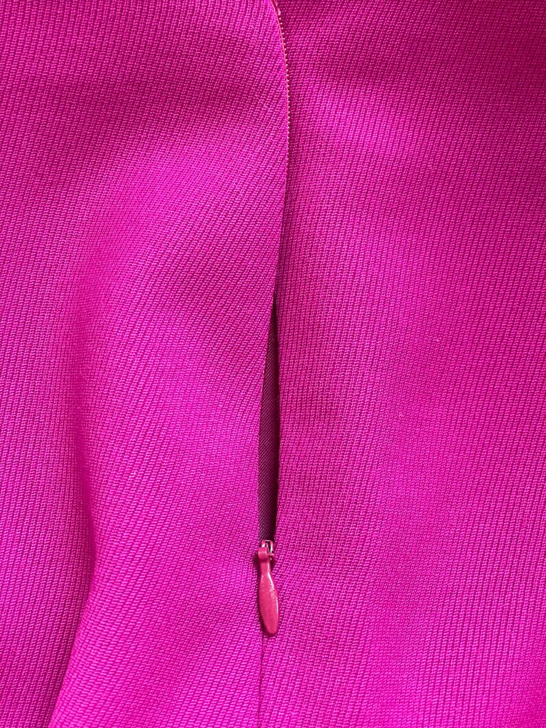 May include: A vibrant magenta fabric surface featuring a concealed zipper closure. The textile has a distinct diagonal weave pattern, and the matching magenta zipper pull is positioned vertically along the seam of the garment material.
