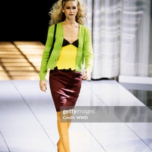 May include: A lime green button-down cardigan worn over a yellow tank top with black lace trim, paired with a dark maroon knee-length skirt, mustard yellow tights, and dark shoes. Light green sunglasses are perched atop blonde hair.