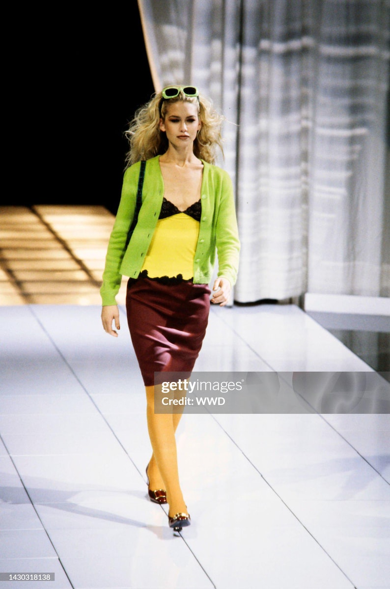 May include: A lime green button-down cardigan worn over a yellow tank top with black lace trim, paired with a dark maroon knee-length skirt, mustard yellow tights, and dark shoes. Light green sunglasses are perched atop blonde hair.