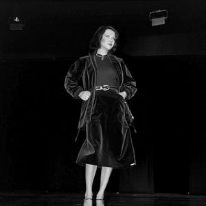 Puede incluir: Un conjunto de chaqueta de terciopelo negro y falda midi a juego con cremallera frontal, mangas largas y cintura entallada con cinturón. El atuendo se combina con una parte superior oscura y un collar sencillo, creando un estilo monocromático.