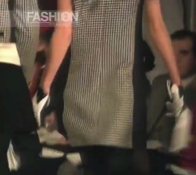 May include: A black and white checkered and striped top with the word "FASHION" in the upper left corner. The top is worn with black pants. The image appears to be from a fashion show.