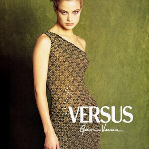 May include: A sleeveless one-shoulder dress featuring a repeating geometric pattern in shades of black, gold, and brown. The text VERSUS Gianni Versace is printed in white on the lower right side of the garment, which has a fitted silhouette.