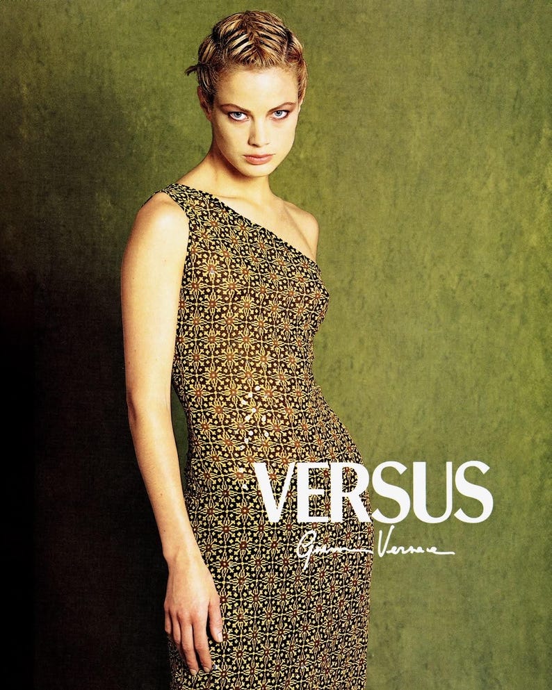 May include: A sleeveless one-shoulder dress featuring a repeating geometric pattern in shades of black, gold, and brown. The text VERSUS Gianni Versace is printed in white on the lower right side of the garment, which has a fitted silhouette.