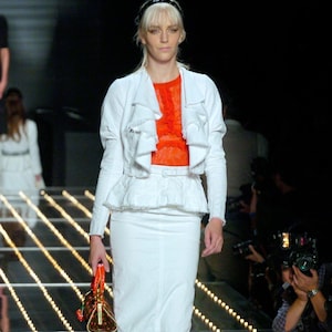 May include: A white jacket with ruffled lapels and a matching peplum skirt with a thin belt, worn over a bright orange lace top. The outfit is paired with a small, structured orange handbag featuring a dark patterned design and gold hardware.