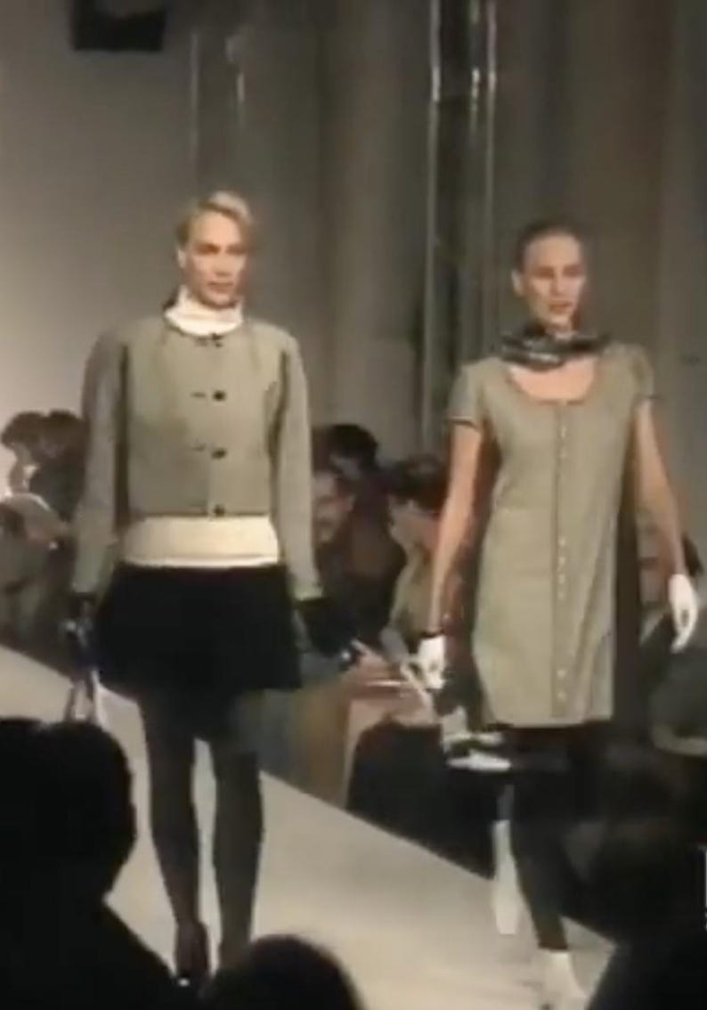 May include: Two models on a runway showcase a fashion collection. The first model wears a light green jacket over a white top and a black skirt. The second model wears a light green dress with a scarf.
