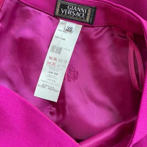 May include: A vibrant magenta skirt featuring a Gianni Versace Couture label and a size 38 tag. The interior is lined with matching magenta fabric and includes a care instruction label detailing 100% wool composition and 100% rayon lining.