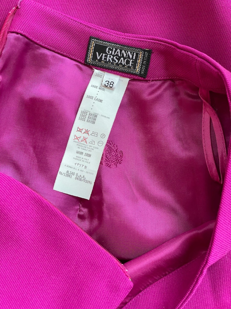 May include: A vibrant magenta skirt featuring a Gianni Versace Couture label and a size 38 tag. The interior is lined with matching magenta fabric and includes a care instruction label detailing 100% wool composition and 100% rayon lining.