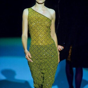 May include: A sleeveless, one-shoulder dress featuring a repeating geometric floral pattern in shades of yellow and black. The fitted garment extends to the mid-calf and is embellished with small, reflective sequins throughout the fabric.