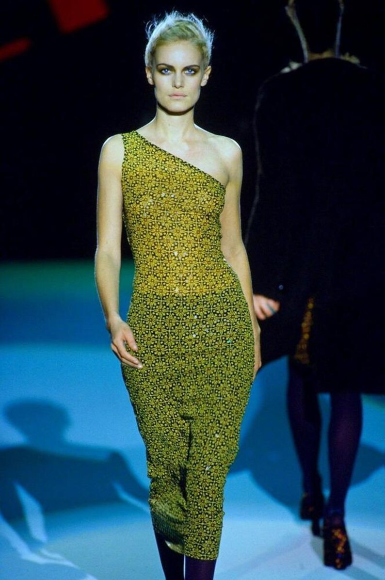 May include: A sleeveless, one-shoulder dress featuring a repeating geometric floral pattern in shades of yellow and black. The fitted garment extends to the mid-calf and is embellished with small, reflective sequins throughout the fabric.