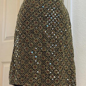 May include: A knee-length skirt featuring a repeating geometric pattern in shades of black, gold, and dark red. The fabric is embellished with small, reflective silver sequins scattered throughout the design, creating a textured appearance.