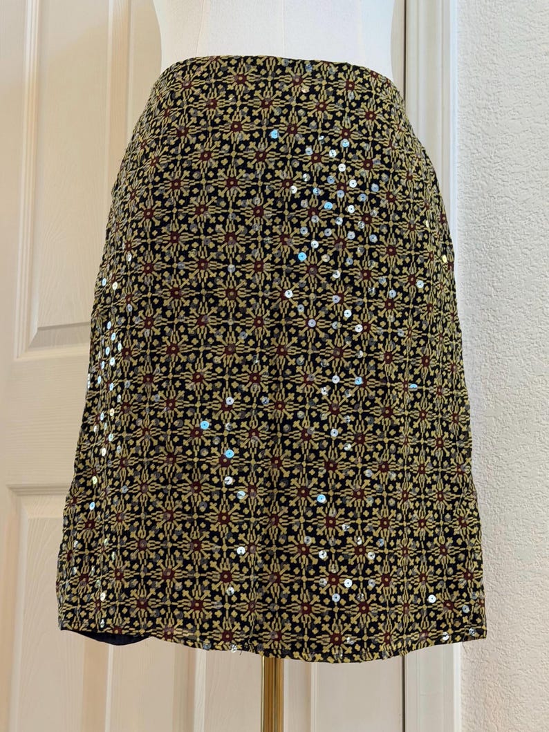 May include: A knee-length skirt featuring a repeating geometric pattern in shades of black, gold, and dark red. The fabric is embellished with small, reflective silver sequins scattered throughout the design, creating a textured appearance.