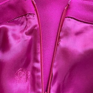 May include: A vibrant magenta garment featuring a central vertical zipper closure and a metal hook-and-eye fastener at the top. The fabric has a smooth, satin-like finish with a repeating tonal embroidered emblem pattern throughout the material.