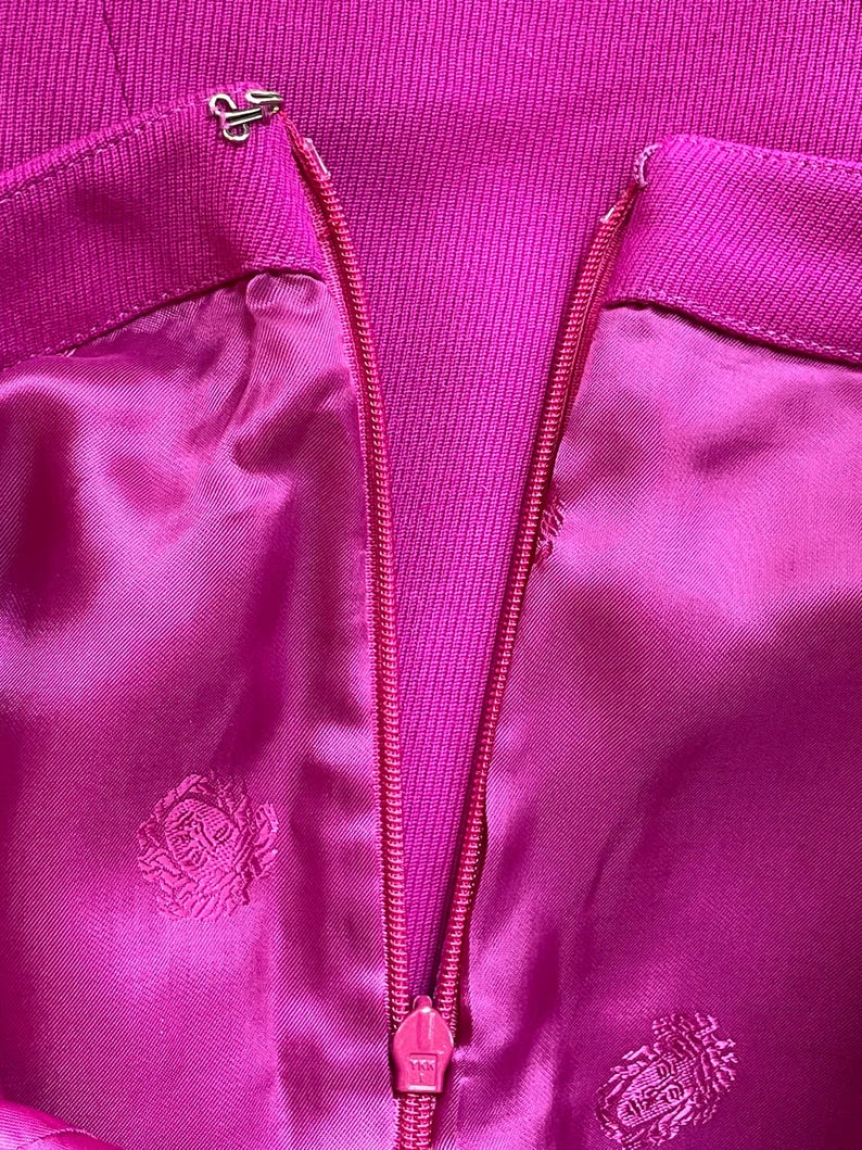 May include: A vibrant magenta garment featuring a central vertical zipper closure and a metal hook-and-eye fastener at the top. The fabric has a smooth, satin-like finish with a repeating tonal embroidered emblem pattern throughout the material.