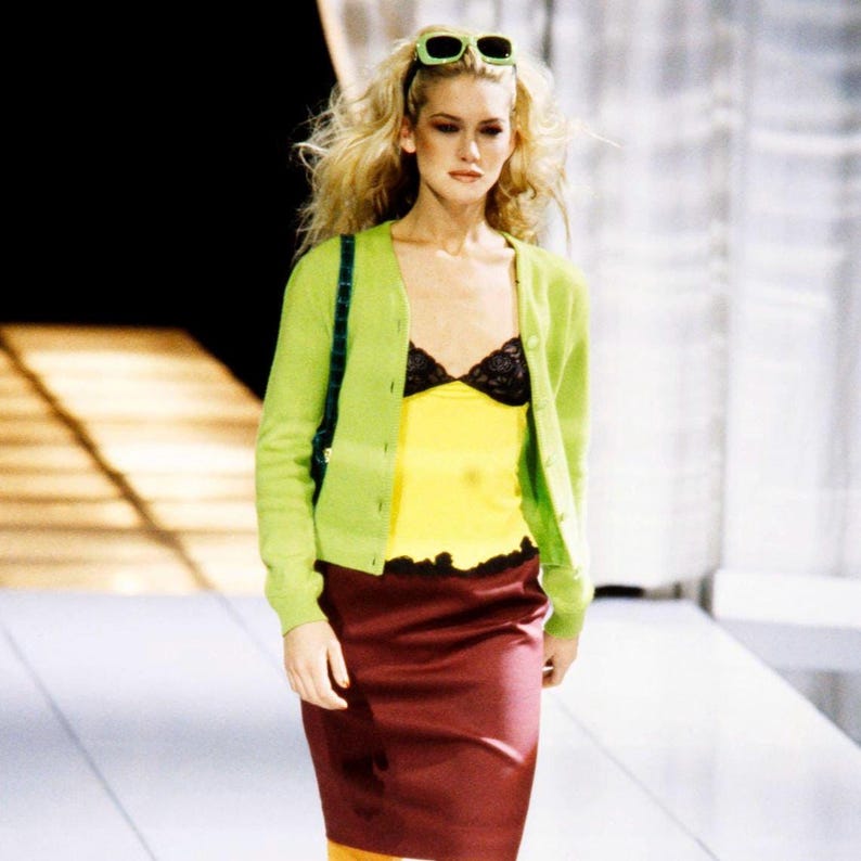 May include: A lime green button-down cardigan worn over a yellow camisole with black lace trim at the neckline and hem. The outfit includes a knee-length maroon skirt and lime green sunglasses resting on top of blonde hair. The ensemble is layered.