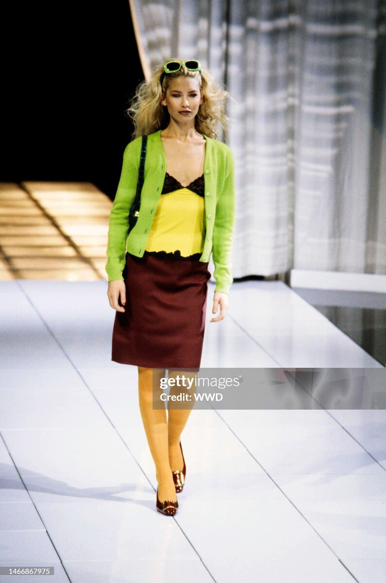 May include: A lime green button-down cardigan worn over a yellow camisole with black lace trim, paired with a knee-length maroon skirt and mustard yellow tights. The outfit includes dark shoes and light green sunglasses worn on the head.