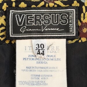 May include: A black clothing label with white text reading VERSUS Gianni Versace Made in Italy, attached to a white care tag with the numbers 30 and 44, manufacturing details, and 100% silk composition, set against a black and yellow patterned fabric.