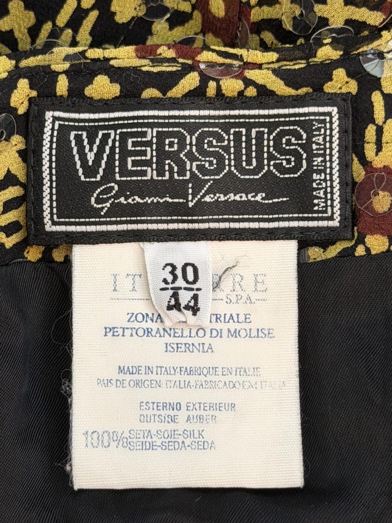 May include: A black clothing label with white text reading VERSUS Gianni Versace Made in Italy, attached to a white care tag with the numbers 30 and 44, manufacturing details, and 100% silk composition, set against a black and yellow patterned fabric.