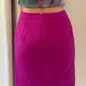 May include: A solid magenta high-waisted skirt featuring a centered vertical seam and a concealed zipper closure at the top. The skirt has a straight-cut silhouette that falls above the knee, constructed from a smooth, opaque fabric with subtle darts.