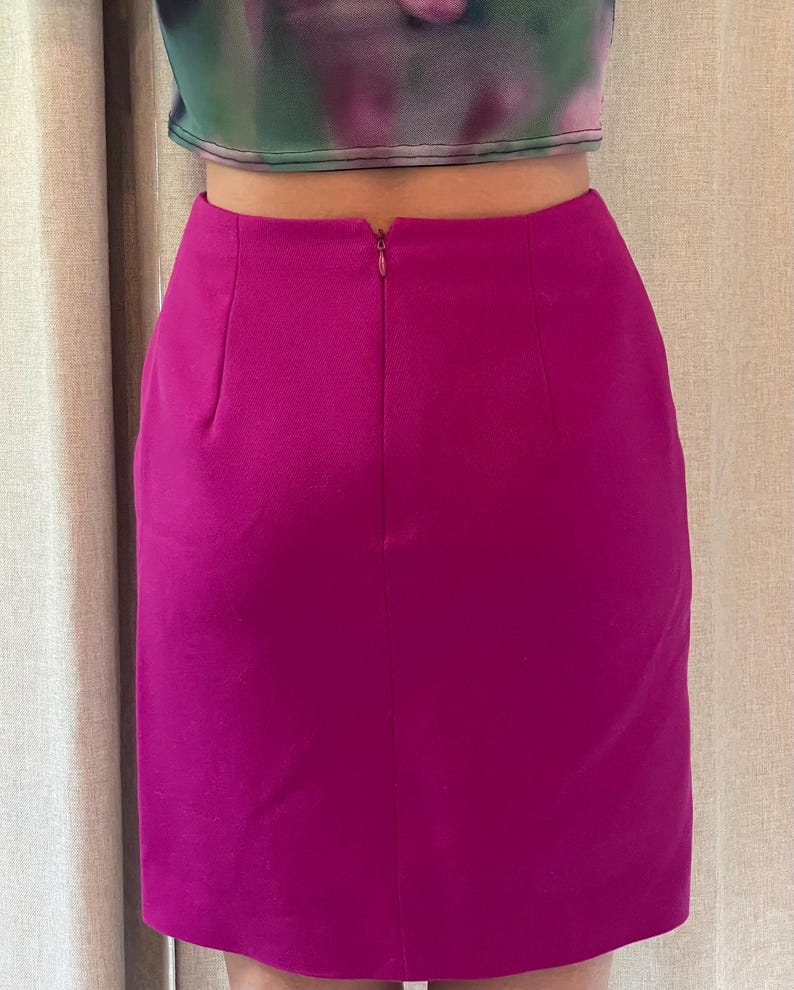 May include: A solid magenta high-waisted skirt featuring a centered vertical seam and a concealed zipper closure at the top. The skirt has a straight-cut silhouette that falls above the knee, constructed from a smooth, opaque fabric with subtle darts.