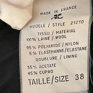 May include: A white fabric tag with black text, featuring details like "MADE IN FRANCE," "100% WOOL," and "SIZE 38." The tag also lists fabric composition and style information. The tag is partially obscured by a white fabric.