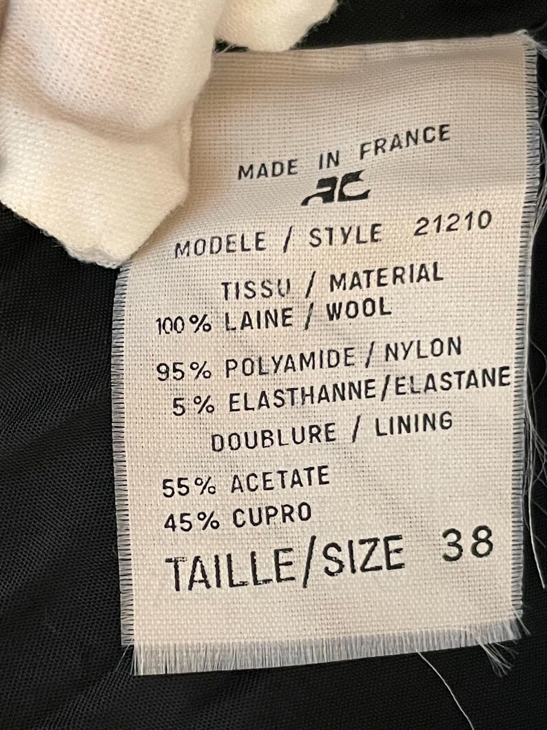 May include: A white fabric tag with black text, featuring details like "MADE IN FRANCE," "100% WOOL," and "SIZE 38." The tag also lists fabric composition and style information. The tag is partially obscured by a white fabric.