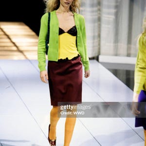 May include: A lime green button-down cardigan worn over a yellow camisole with black lace trim, paired with a knee-length maroon skirt, mustard yellow tights, and dark brown shoes with gold buckles. A dark green shoulder bag is carried on the side.
