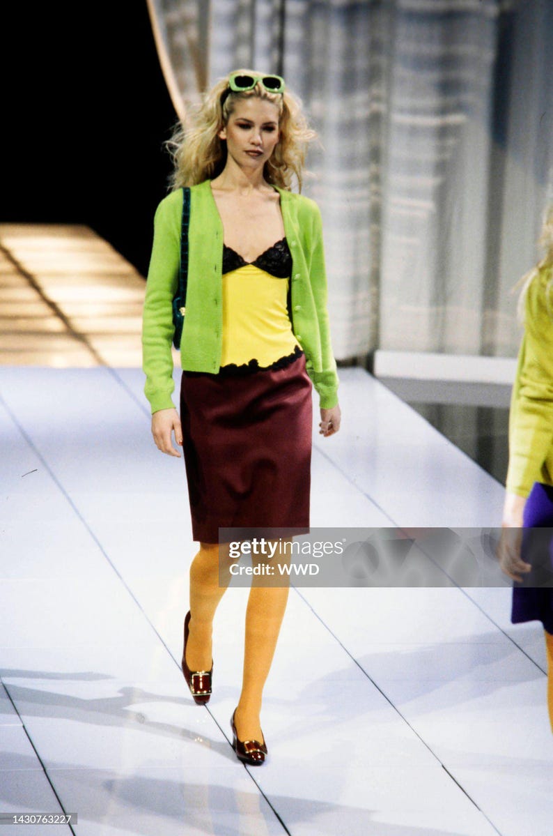 May include: A lime green button-down cardigan worn over a yellow camisole with black lace trim, paired with a knee-length maroon skirt, mustard yellow tights, and dark brown shoes with gold buckles. A dark green shoulder bag is carried on the side.