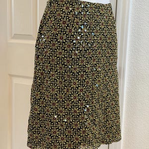 May include: A knee-length skirt featuring a black base with a repeating geometric pattern in yellow and dark red. The fabric is embellished with scattered silver sequins that catch the light, creating a textured appearance across the surface.