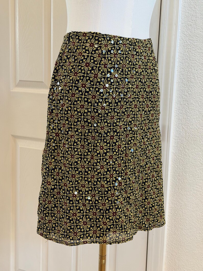 May include: A knee-length skirt featuring a black base with a repeating geometric pattern in yellow and dark red. The fabric is embellished with scattered silver sequins that catch the light, creating a textured appearance across the surface.