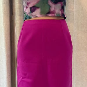 May include: A solid magenta knee-length skirt with a straight-cut silhouette and a smooth, matte finish. The skirt features a clean, minimalist design with no visible pockets, zippers, or embellishments, providing a simple and structured appearance.