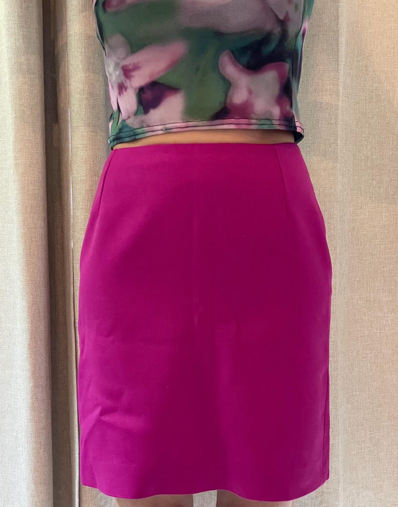 May include: A solid magenta knee-length skirt with a straight-cut silhouette and a smooth, matte finish. The skirt features a clean, minimalist design with no visible pockets, zippers, or embellishments, providing a simple and structured appearance.