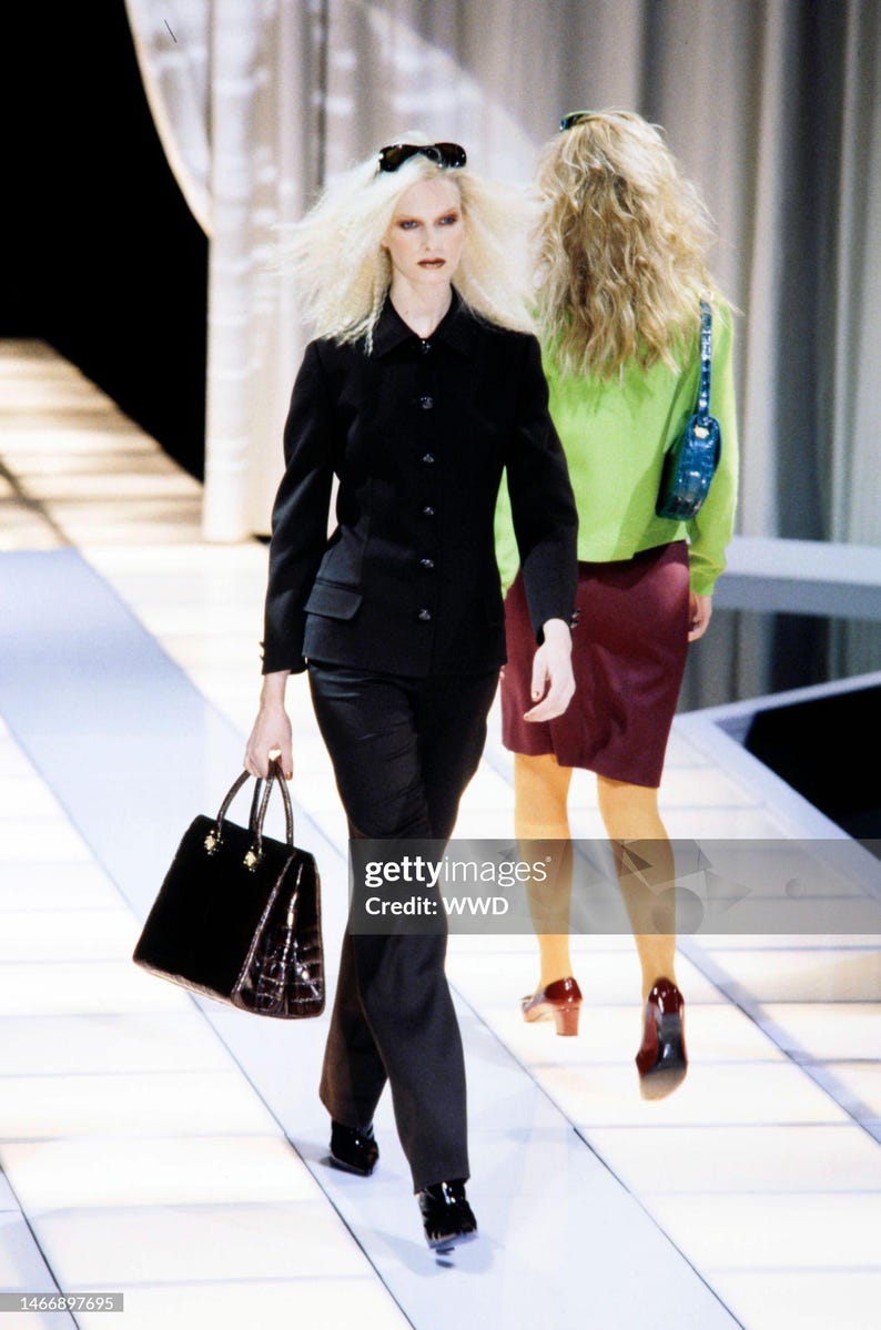 May include: A black button-down blazer with matching black trousers and a black structured handbag. The outfit is paired with black shoes and dark sunglasses worn on the head. A second outfit features a lime green jacket, a maroon skirt, and yellow tights.