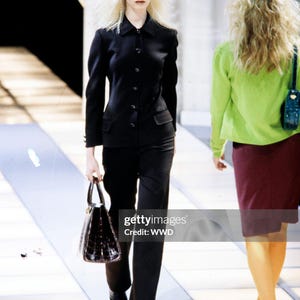 May include: A black long-sleeved button-down jacket with a collar and two front pockets, paired with matching black trousers. The outfit is accessorized with a dark brown structured handbag and black pointed-toe shoes.