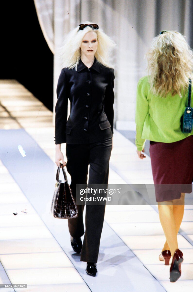 May include: A black long-sleeved button-down jacket with a collar and two front pockets, paired with matching black trousers. The outfit is accessorized with a dark brown structured handbag and black pointed-toe shoes.