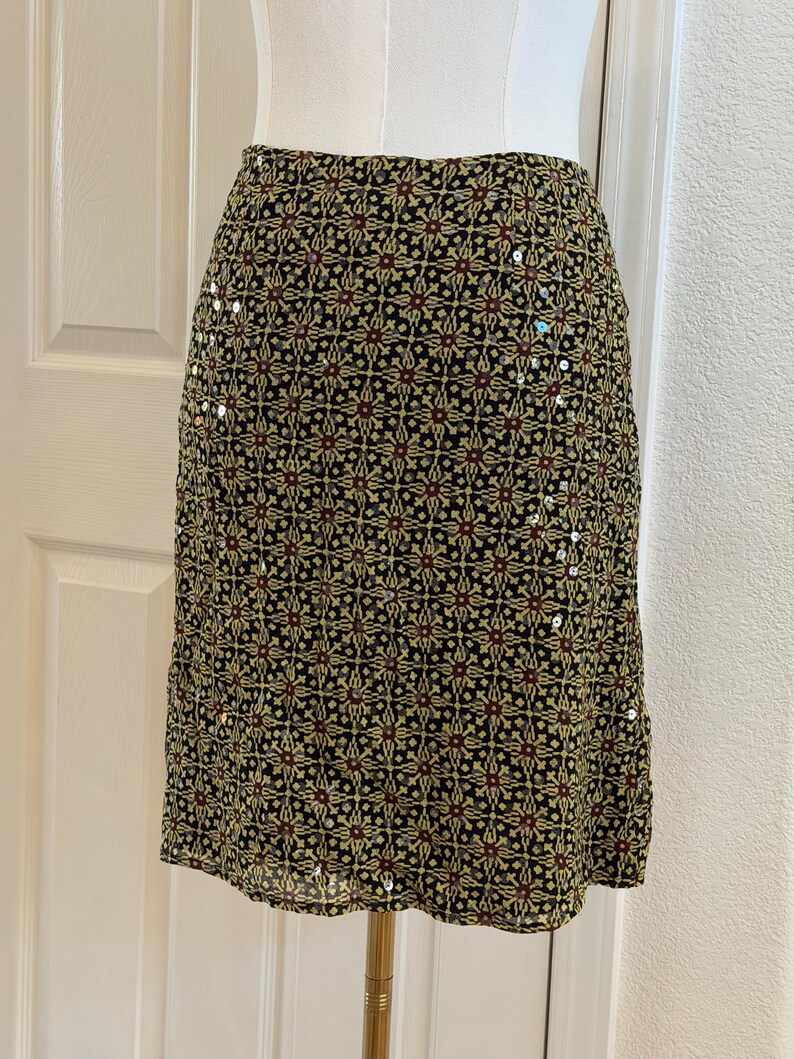 May include: A knee-length skirt featuring a black base with a repeating geometric pattern in yellow and dark red. The fabric is embellished with small, scattered silver sequins that catch the light, creating a subtle shimmer across the surface.