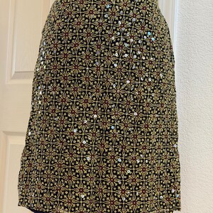 May include: A knee-length skirt featuring a black base with a repeating geometric pattern in gold and dark red. The fabric is embellished with scattered silver sequins that catch the light, creating a textured appearance across the entire surface.