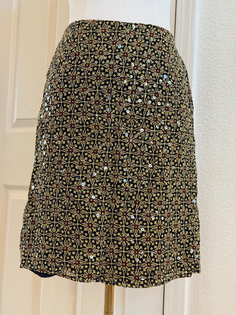 May include: A knee-length skirt featuring a black base with a repeating geometric pattern in gold and dark red. The fabric is embellished with scattered silver sequins that catch the light, creating a textured appearance across the entire surface.