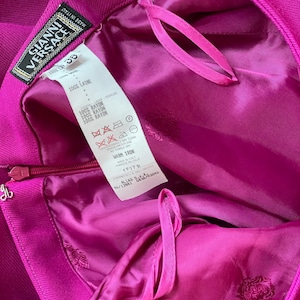 May include: A bright magenta garment featuring a Gianni Versace label and a white care tag with text including 100% LAINE and 100% RAYON. The interior is lined with matching magenta fabric, and a zipper closure is visible on the side.