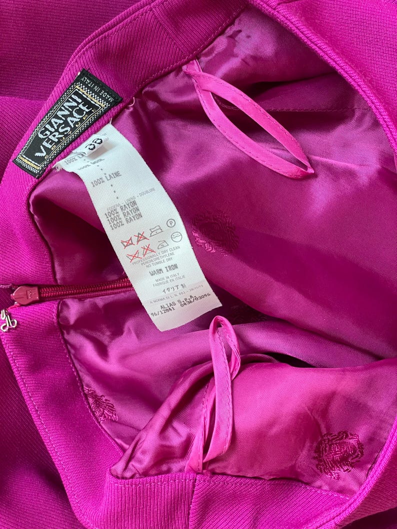 May include: A bright magenta garment featuring a Gianni Versace label and a white care tag with text including 100% LAINE and 100% RAYON. The interior is lined with matching magenta fabric, and a zipper closure is visible on the side.