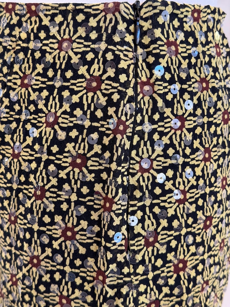 May include: A black skirt featuring a repeating geometric pattern in yellow and dark red, embellished with small, clear, circular sequins throughout the fabric. The garment includes a vertical black zipper closure centered along the back seam.