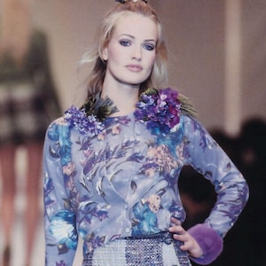 May include: A long-sleeved button-down cardigan featuring a blue and purple floral print. The garment is adorned with clusters of purple and green fabric flowers on each shoulder and includes a solid purple faux fur cuff on the left sleeve.
