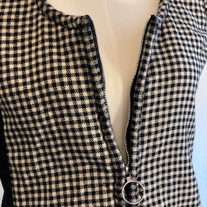 May include: A black and white checkered vest with a silver zipper. The zipper has a circular pull. The vest is displayed on a mannequin. The fabric has a small, repeating pattern.