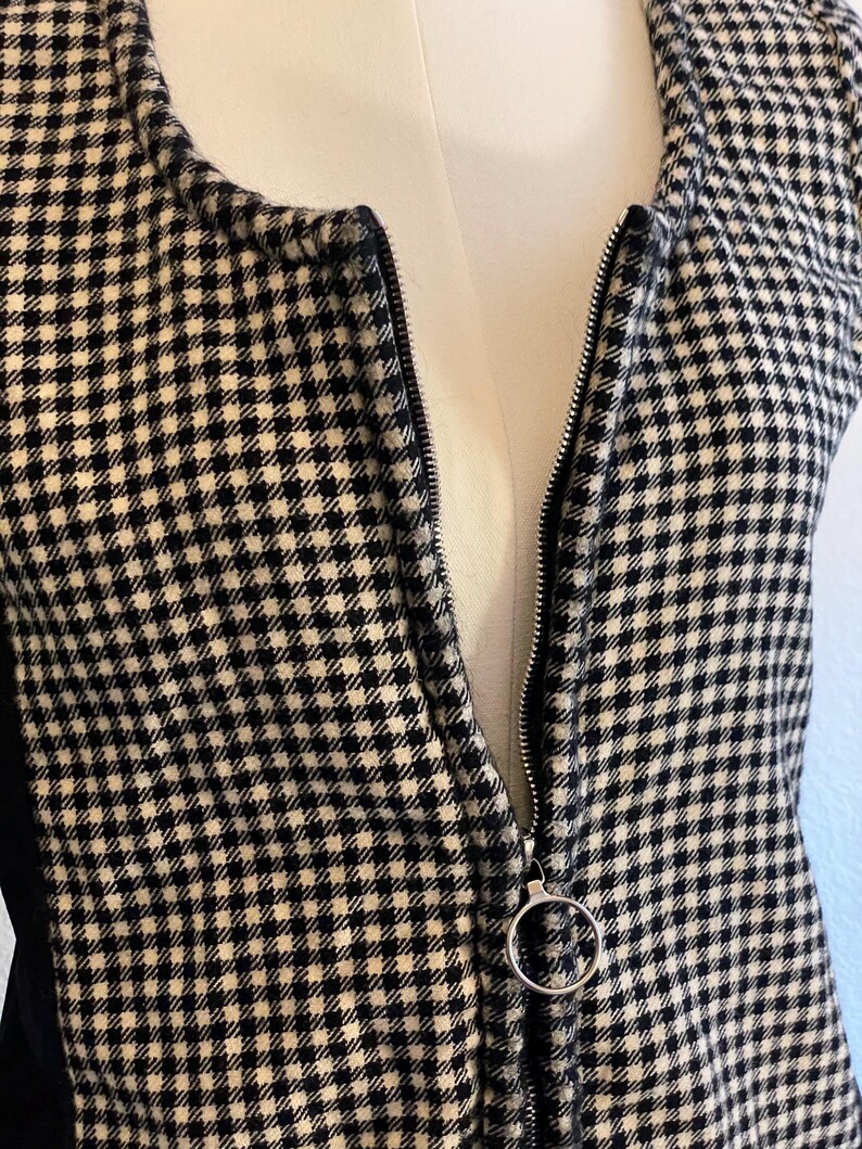May include: A black and white checkered vest with a silver zipper. The zipper has a circular pull. The vest is displayed on a mannequin. The fabric has a small, repeating pattern.