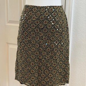 May include: A knee-length skirt featuring a black base with a repeating geometric pattern in shades of yellow, red, and green. The fabric is embellished with small, scattered silver sequins that catch the light across the surface of the garment.
